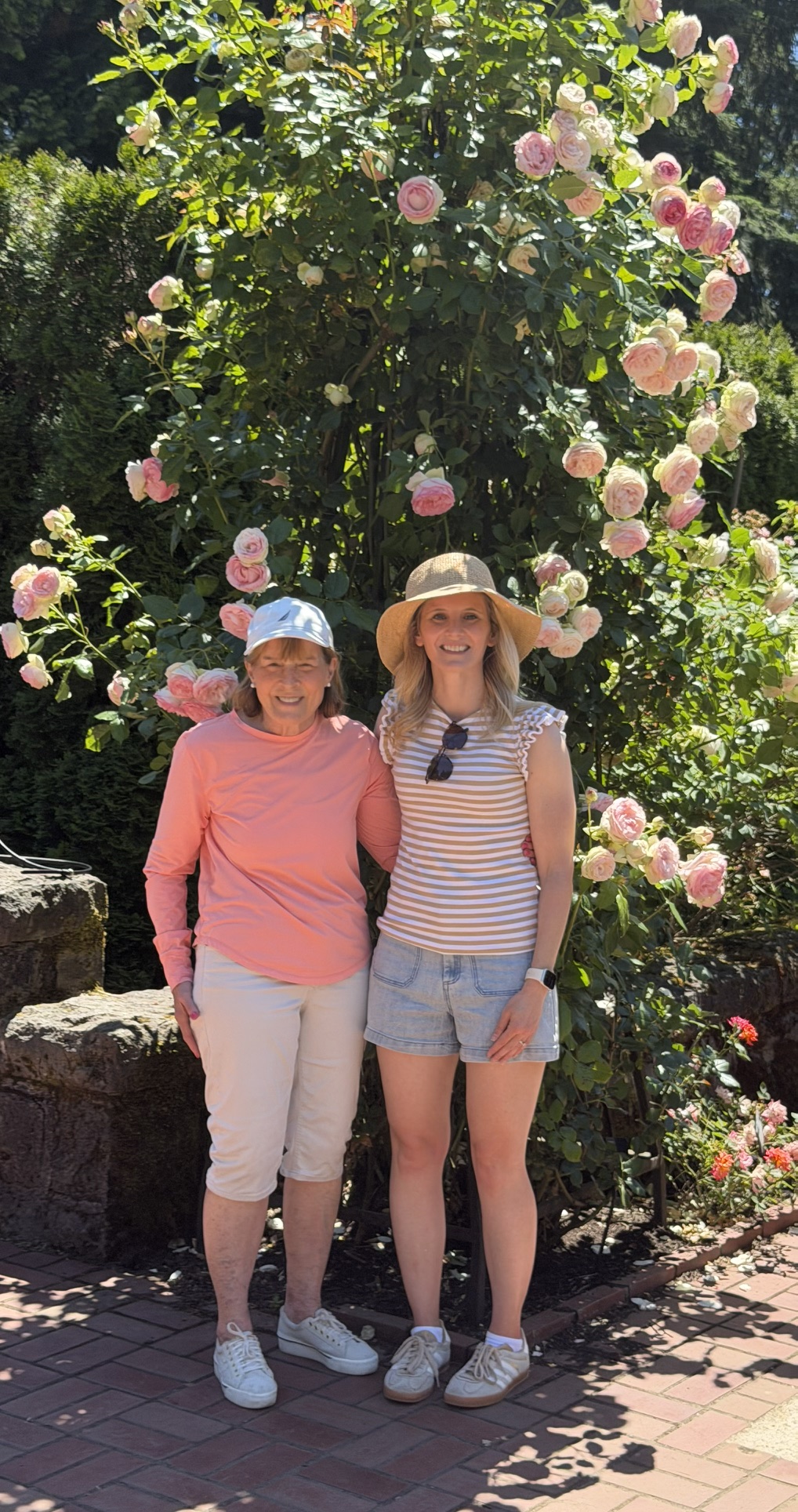 Two women standing in front of a tall Eden climbing rose covered in blush and cream blooms at the Portland International Rose Test Garden.
